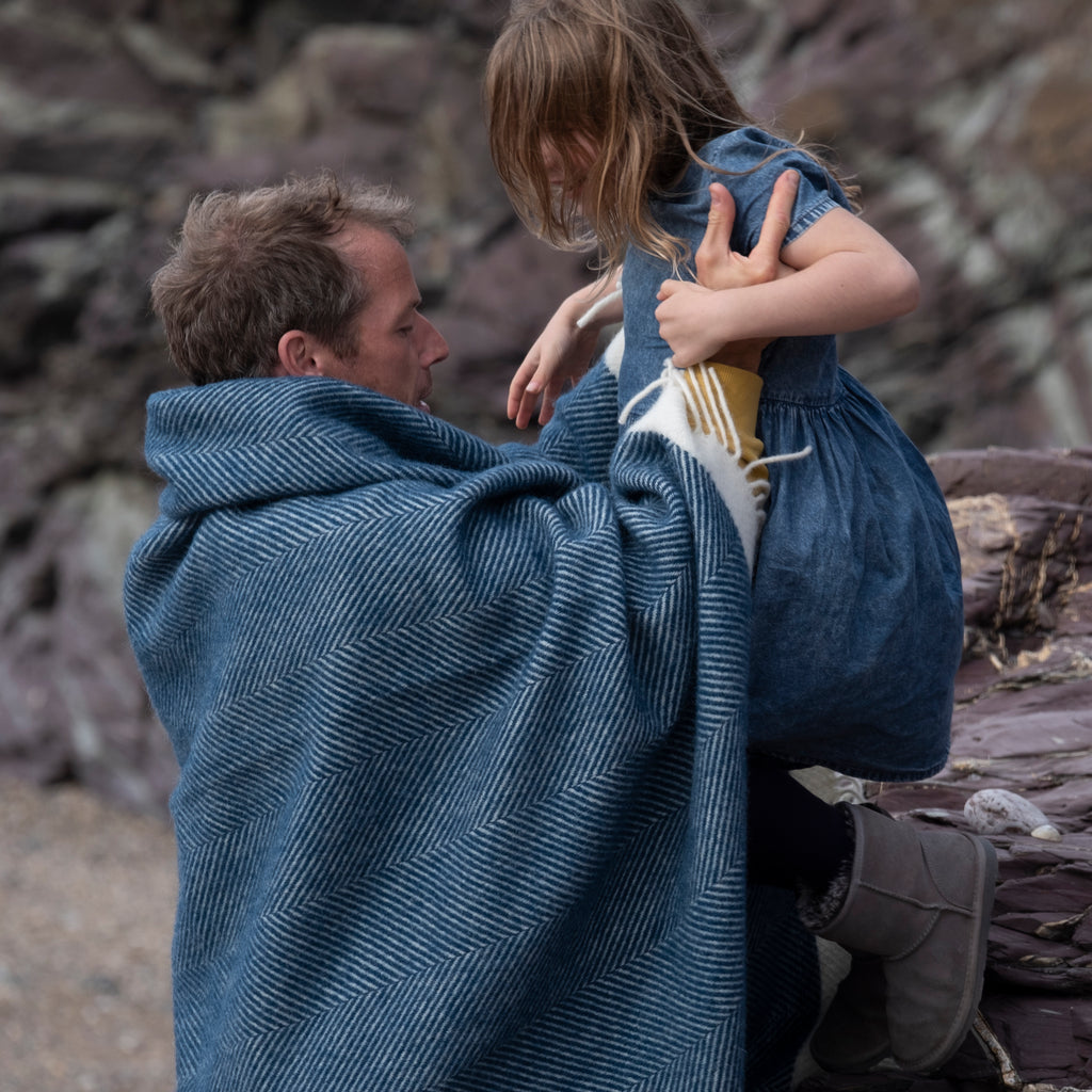 man with navy wool blanket wrapped around shoulders holding child on beach